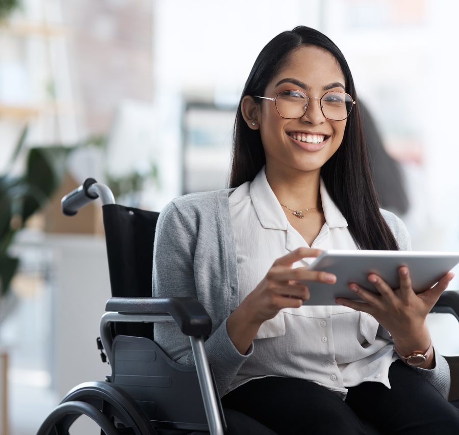 A smiling woman seated in her wheelchair and using a tablet in a bright office